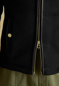 Čierna fleecová bunda so zlatým zipsom, čiastočne otvorená, pod ňou sú vidieť olivovo zelené nohavice a malý zlatý cvok na vrecku.