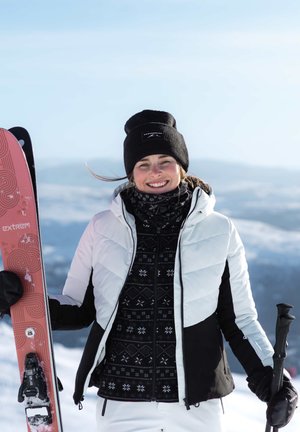 Femme souriante en veste d'hiver et chapeau tenant des skis roses et un bâton de ski avec un paysage montagneux enneigé en arrière-plan.