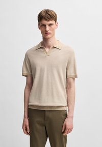 Polo tricotat bej cu decolteu în V, mâneci scurte și tiv ribbelat. Material texturat, asortat cu pantaloni verzi măslinii.