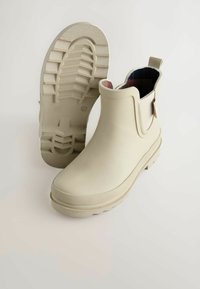 Bottes chelsea en caoutchouc beige clair avec semelle texturée, languette de traction et doublure intérieure à carreaux, conçues pour un usage imperméable.