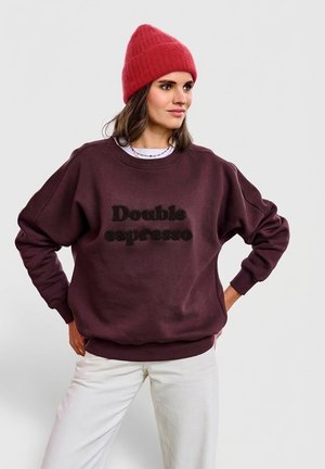 Bordowa bluza z napisem „Double espresso”, oversize, z okrągłym dekoltem i ściągaczami na rękawach. Noszona z jasnymi spodniami i czerwoną czapką beanie.