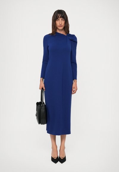 Donna che indossa un vestito blu royal a maniche lunghe e lunghezza midi con dettagli sulle spalle, tiene una borsa nera e indossa tacchi a punta neri.