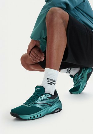 Osoba w turkusowych butach do biegania Reebok, białych skarpetkach Reebok, czarnych szortach i turkusowej kurtce, kucająca na białym tle.