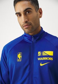 Mėlyna sportinė striukė, pagaminta iš lygaus audinio, su NBA logotipu, užrašu "WARRIORS" geltona spalva ir stilizuota tilto grafiką. Matomas Nike logotipas.
