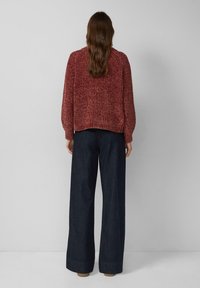 Roter strukturiert gestrickter Pullover mit lockerem Schnitt, kombiniert mit weit geschnittenen dunkelblauen Jeanshosen, von hinten vor hellem Hintergrund gesehen.