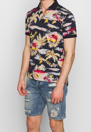 Mężczyzna w czarnej koszulce polo z tropikalnym nadrukiem palm i czerwonym napisem, w połączeniu z jasnoniebieskimi podartymi szortami dżinsowymi.