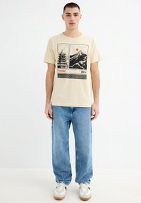Camiseta de algodón beige con un estampado gráfico de montañas y una pagoda, combinada con jeans de denim azul sueltos y zapatillas blancas.
