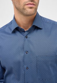 Camicia blu a fantasia con colletto button-down, caratterizzata da piccoli punti bianchi, bottoni marroni a contrasto e una tasca sul petto.