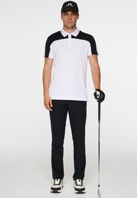 Herrgolfkläder bestående av en vit pikétröja med svarta accenter, svarta byxor och sneakers, som håller i en golvklubba.