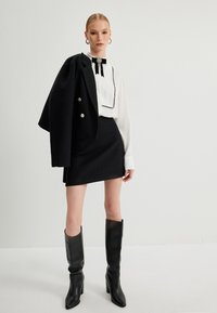 Schwarzer Blazer mit silbernen Knöpfen, weiße Bluse mit schwarzem Saum und Schleife, strukturierter schwarzer Minirock und kniehohe schwarze Stiefel.