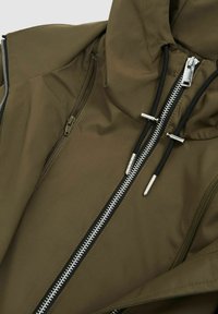 Veste vert olive avec un col haut, plusieurs poches zippées et des détails de fermeture éclair blancs contrastants avec des tirettes noires. Fini lisse et mat.