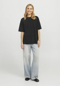 Černé oversized tričko s krátkými rukávy, kombinováno se světle vybledlými širokými džíny. Model má na sobě černé slip-on boty. Neutrální pozadí.