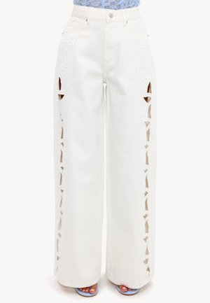 Personne portant un pantalon blanc taille haute à jambes larges avec des motifs découpés verticaux de chaque côté, associé à des chaussures bleues ouvertes.
