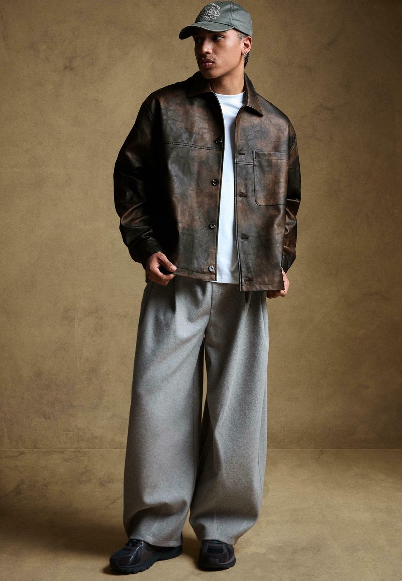 Barna fényes bőr dzseki gombos záródással, alatta fehér póló, szürke bő, széles lábú nadrág és fekete sportcipő. Szürke sapka logóval.