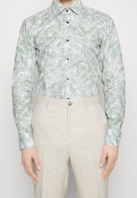 Langarmshirt van lichtgewicht stof met een groen palmenbladpatroon op een lichte achtergrond; gecombineerd met beige linnen broeken.