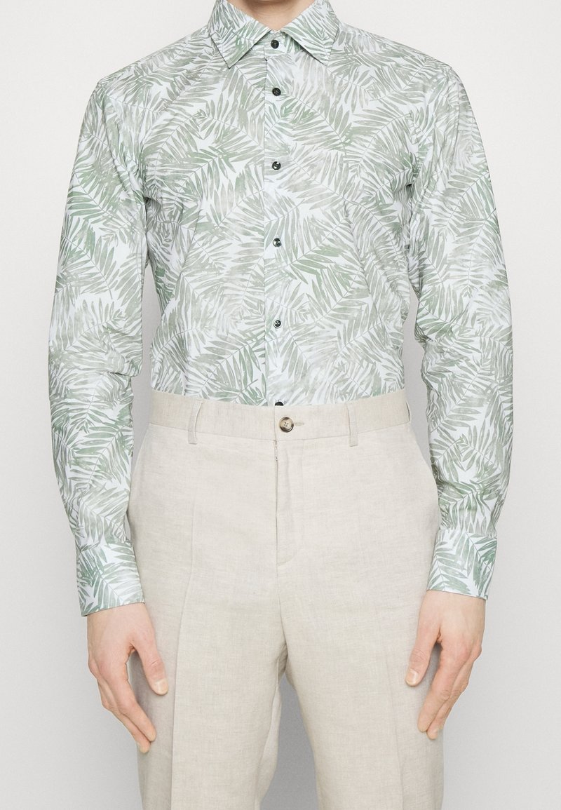 Langarmshirt van lichtgewicht stof met een groen palmenbladpatroon op een lichte achtergrond; gecombineerd met beige linnen broeken.