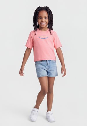 Niña sonriente con el cabello trenzado que lleva una camiseta rosa de Patagonia, pantalones cortos de mezclilla y zapatillas blancas, de pie frente a un fondo sencillo.