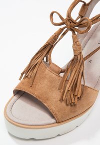 Sandale ouverte à bout ouvert en daim marron avec semelle blanche et pompons décoratifs noués en nœud sur les brides avant.