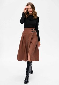 Vrouw in een zwarte lange mouwtop, een bruine midi-rok met een zijsplit en gespsdetails, en zwarte kniehoge laarzen die loopt met één hand bij haar gezicht.
