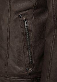Nahaufnahme einer braunen Lederjacke, die eine seitliche Reißverschlusstasche, eine strukturierte Oberfläche und detaillierte Nähte mit einem glatten, matten Finish zeigt.