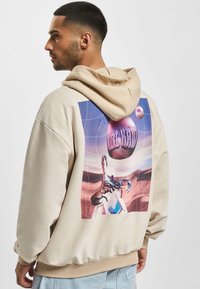 Beige hoodie med et stort grafiktryk, der viser en krabbe og en surrealistisk baggrund. Blødt materiale, afslappet pasform med en justerbar hætte.