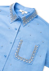 Hemelsblauw shirt met korte mouwen van een gestructureerde stof, versierd met metalen studs en parels op de kraag en de zak. Twee knopen zichtbaar.