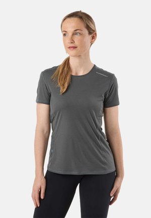 Kvinde med lysebrunt hestehale iført en grå kortærmet sportstrøje og sorte leggings, stående mod en ensfarvet hvid baggrund.