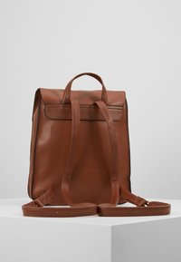 Ruskea nahkainen reppu suorakulmaisella muotoilulla, yläkahvalla, säädettävillä olkahihnoilla ja yksinkertaisella, sileällä pinnalla. Ei näkyviä metalliosia tai kuvioita.