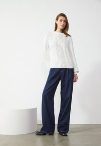 Hvit strikket genser med kabelmønster og ribbestrikket kant, kombinert med vide, mørkeblå denimbukser. Modellen står foran en hvit bakgrunn.