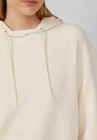 Cremfarbener Hoodie aus weichem Material, mit einer Kordel am Kragen und einer lässigen Passform. Glatte Textur mit minimalen Details.