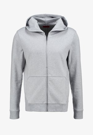 Jasnoszara bluza na zamek w stylu hoodie noszona na czarnej koszulce, w połączeniu z ciemnymi dżinsami i czarnymi butami wsuwanymi. Gładki materiał, luźny krój, minimalistyczny design.