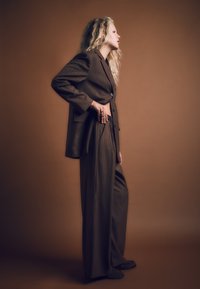 Hnedý pruhovaný oblek s nohavicami so širokými nohavicami a strihaným sako. Materiál vyzerá textúrovaný, s viditeľnými gombíkmi a uvoľneným strihom.