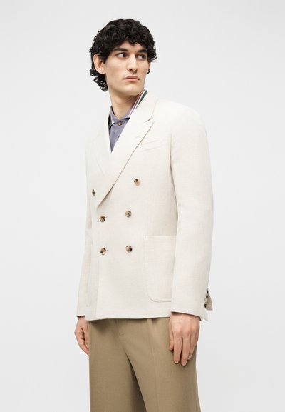 Homme portant un blazer crème à double boutonnage avec des boutons beige clair par-dessus une chemise violette et un pantalon beige, regardant sur le côté.
