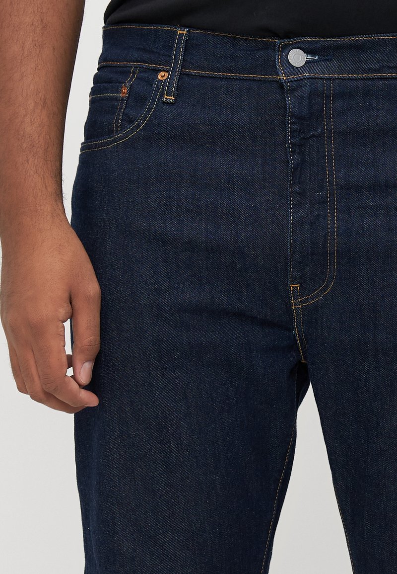 Nahaufnahme einer Person, die dunkelblaue Jeans mit sichtbaren Nähten trägt, während eine Hand entspannt an ihrer Seite gegen einen einfarbigen Hintergrund liegt.
