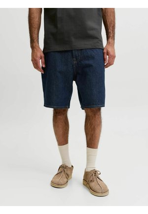 Hombre con pantalones cortos vaqueros azul oscuro, zapatos de ante beige con cordones y calcetines blancos hasta el tobillo, de pie sobre un fondo claro y liso.