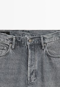 Detailní záběr na šedý opasek u riflí z denimu s poutky na pásek, kovovým knoflíkem, zipem a předními kapsami.