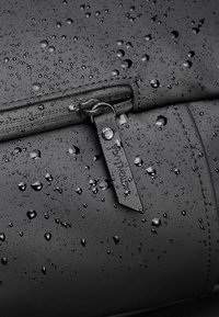 Sac noir résistant à l'eau avec une texture lisse, présentant des gouttes d'eau, une fermeture éclair et une étiquette avec une marque gravée.