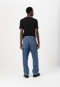Chemise noire à manches courtes associée à un pantalon cargo en denim bleu ample, doté de multiples poches et d'une coupe décontractée, porté avec des chaussures noires.