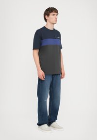 Marineblå og grå kortermet T-skjorte med en horisontal blå stripe, kombinert med lyseblå jeans og hvite joggesko på en ensfarget bakgrunn.