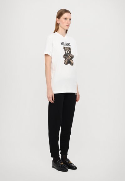 Hvit T-skjorte med grafikk av svart bjørn og "Moschino"-tekst, kombinert med svarte joggebukser og svarte loffer. Mykt bomullsmateriale.