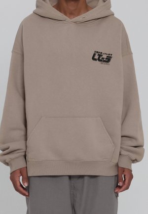 Personne portant un hoodie beige avec un texte japonais noir et un logo sur le côté gauche de la poitrine, associé à un pantalon gris, les mains détendues le long du corps.