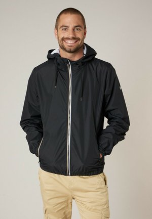 Homme souriant aux cheveux courts portant une veste noire à capuche et un pantalon cargo beige, debout les mains dans les poches devant un arrière-plan uni.