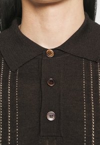 Hnědá pletená polo tričko s límcem, obsahující tři knoflíkové uzávěry a vertikální pletené vzory s drobnými otevřenými detaily.