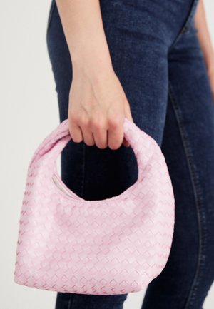 Hand holding a small pink woven handbag with a rounded handle, worn with dark blue jeans against a plain background.