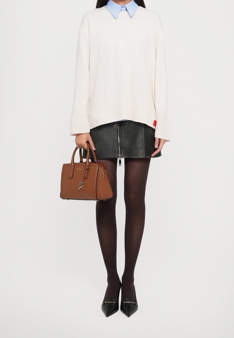 Personne portant un pull blanc sur une chemise bleue à col, une jupe noire, des collants noirs et des chaussures noires pointues, tenant un sac à main en cuir marron.