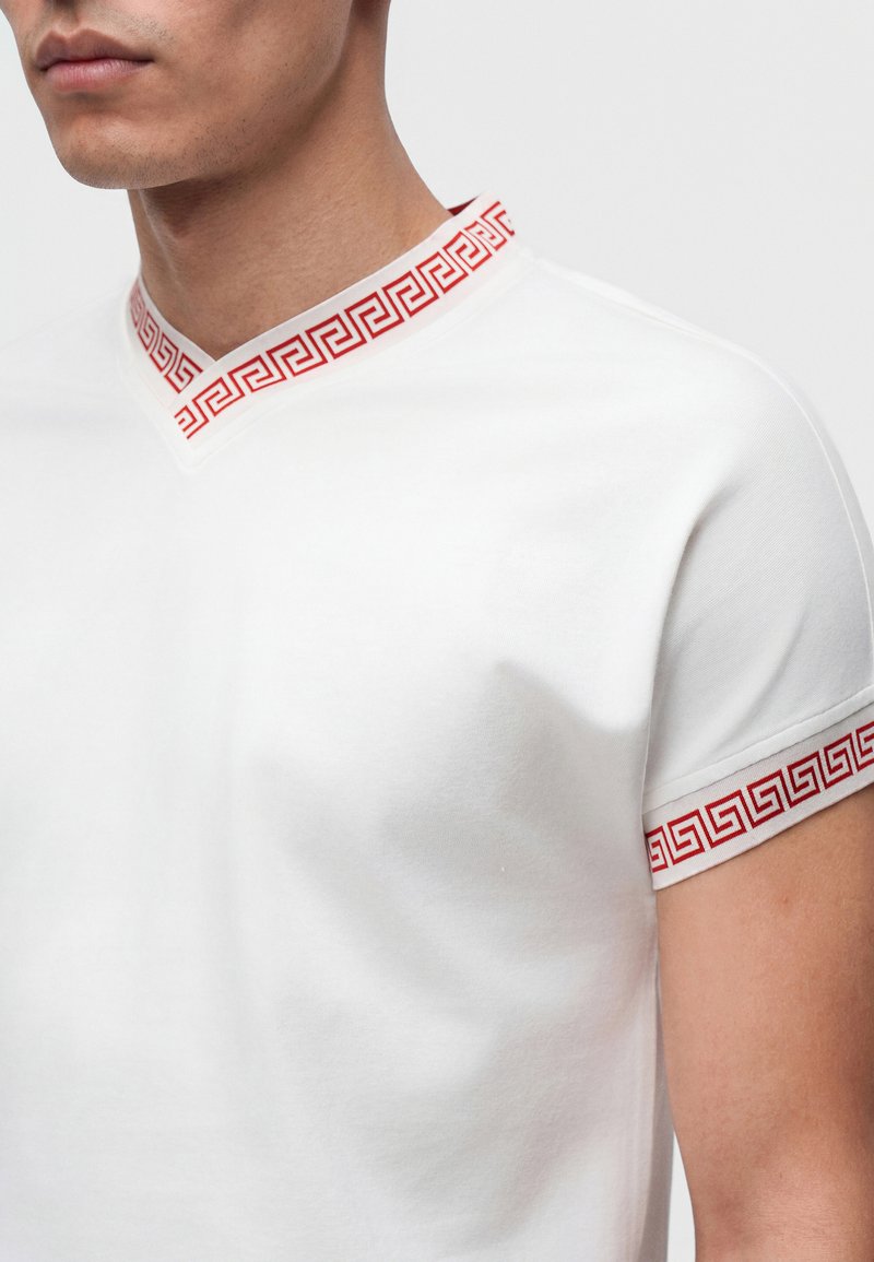 Gros plan d'une personne portant une chemise blanche avec une bordure à motif géométrique rouge sur le col et les bords des manches.
