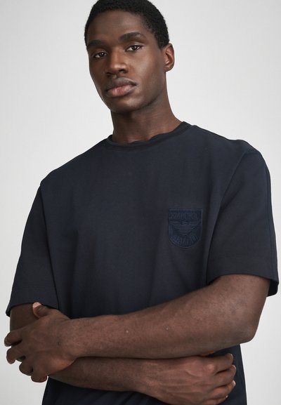 Mørkeblå bomull T-skjorte med rund hals og korte ermer; har et teksturert emblem på venstre bryst. Glatt stoff, avslappet passform.