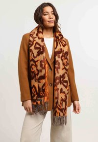 Sciarpa con stampa animale marrone realizzata in materiale morbido, con accenti arancioni e dettagli frangiati, indossata sopra un cappotto marrone e abbinata a pantaloni beige.