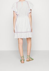 Vestido blanco de tirantes acampanados y espalda con rendija, con un dobladillo con volantes y acentos en lavanda. Combinado con sandalias verdes de tiras.