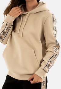 Felpa beige con tasca a marsupio, cappuccio con cordino e nastro con logo nero a strisce lungo le maniche. Tessuto morbido con una vestibilità comoda.